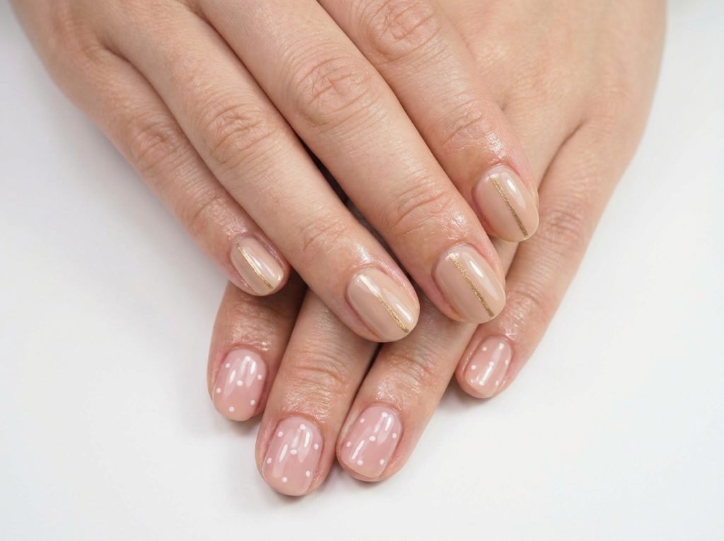 "Close-up of two hands with freshly painted minimalist nail art: one hand showing a nude base with a single thin gold stripe down the center of each nail, the other with small white dots on a blush pink base β clean white background, macro photography"