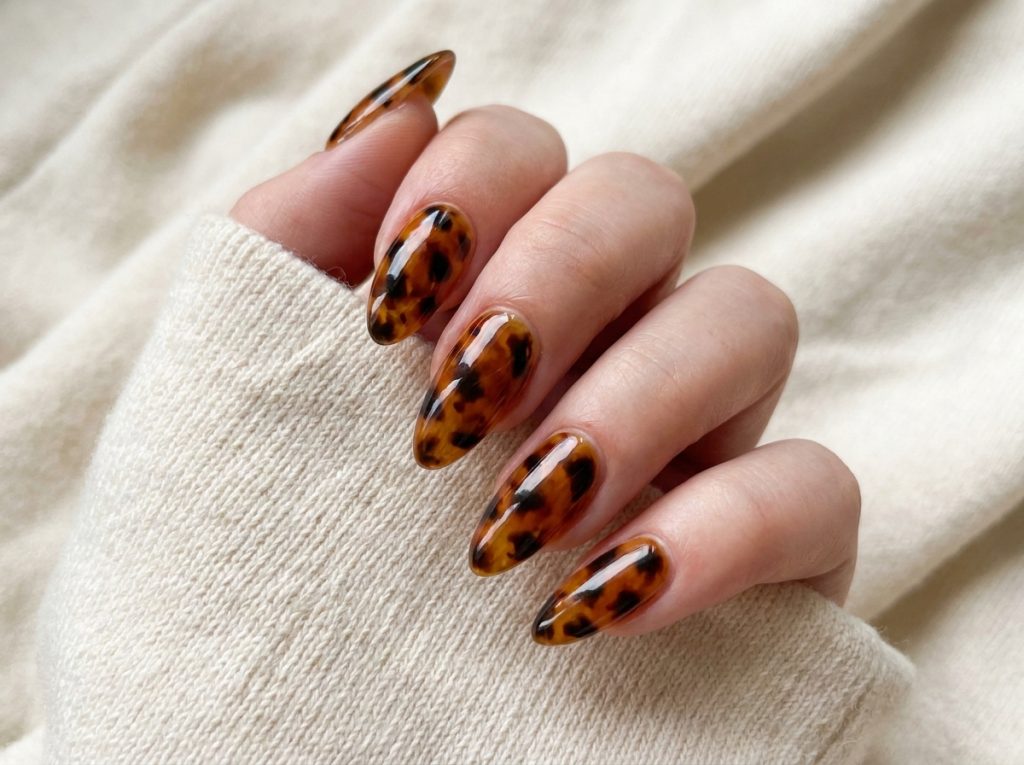 "Close-up of tortoiseshell nail art on almond-shaped nails β warm amber base with irregular dark brown and black patches, high-gloss finish, elegant hand pose on cream fabric background"